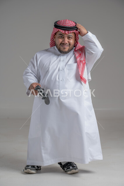 Spending free time playing PlayStation entertainment games, gestures of happiness and pleasure, entertaining modern technologies, portrait of a young Saudi Gulf dwarf with special needs wearing traditional thobe and shemagh holding a joystick in his hand, full-length body image, gray background
