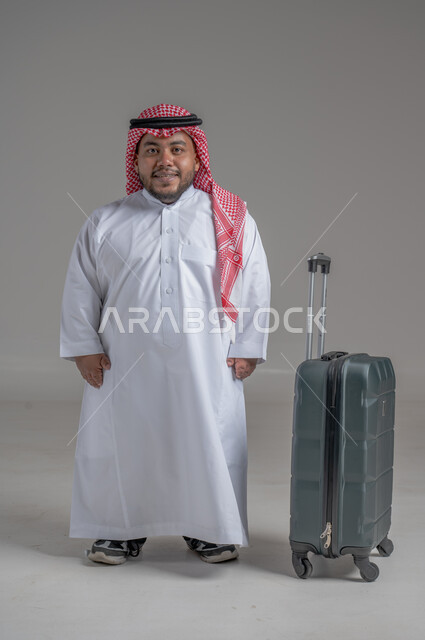 Communicating with family and friends, making phone calls via mobile, portrait of a smiling Saudi Arabian young man, Gulf dwarf, wearing traditional thobe and shemagh, holding a mobile phone in his hand and a travel bag next to him, direct booking via tourism applications and programs, full-length body image, gray background