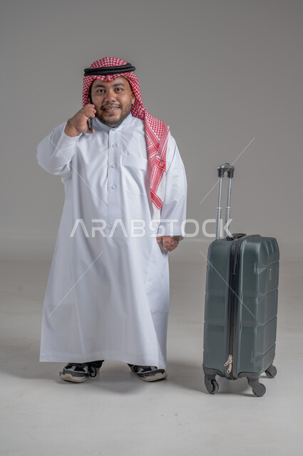 التواصل مع الأهل والأصدقاء, إجراء مكالمات هاتفية عبر الجوال, بورتريه لشاب عربي سعودي قزم خليجي مبتسم يرتدي الثوب التقليدي والشماغ يمسك بيده الهاتف المحمول وبجانبه حقيبة السفر, حجز مباشر عبر تطبيقات وبرامج السياحة, صورة الجسم بالطول الكامل, خلفية رمادية