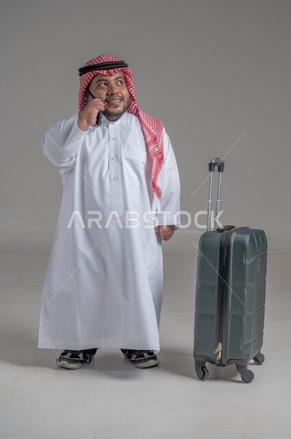 Communicating with family and friends, making phone calls via mobile, portrait of a smiling Saudi Arabian young man, Gulf dwarf, wearing traditional thobe and shemagh, holding a mobile phone in his hand and a travel bag next to him, direct booking via tourism applications and programs, full-length body image, gray background