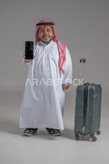 التواصل مع الأهل والأصدقاء, إجراء مكالمات هاتفية عبر الجوال, بورتريه لشاب عربي سعودي قزم خليجي مبتسم يرتدي الثوب التقليدي والشماغ يمسك بيده الهاتف المحمول وبجانبه حقيبة السفر, حجز مباشر عبر تطبيقات وبرامج السياحة, صورة الجسم بالطول الكامل, خلفية رمادية