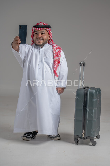 التواصل مع الأهل والأصدقاء, إجراء مكالمات هاتفية عبر الجوال, بورتريه لشاب عربي سعودي قزم خليجي مبتسم يرتدي الثوب التقليدي والشماغ يمسك بيده الهاتف المحمول وبجانبه حقيبة السفر, حجز مباشر عبر تطبيقات وبرامج السياحة, صورة الجسم بالطول الكامل, خلفية رمادية