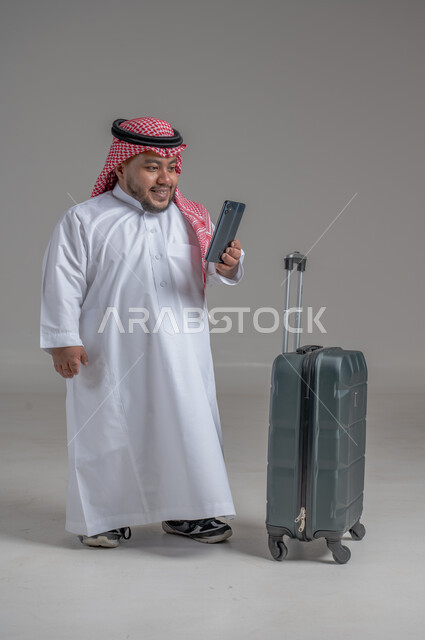 Communicating with family and friends, making phone calls via mobile, portrait of a smiling Saudi Arabian young man, Gulf dwarf, wearing traditional thobe and shemagh, holding a mobile phone in his hand and a travel bag next to him, direct booking via tourism applications and programs, full-length body image, gray background