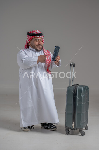 التواصل مع الأهل والأصدقاء, إجراء مكالمات هاتفية عبر الجوال, بورتريه لشاب عربي سعودي قزم خليجي مبتسم يرتدي الثوب التقليدي والشماغ يمسك بيده الهاتف المحمول وبجانبه حقيبة السفر, حجز مباشر عبر تطبيقات وبرامج السياحة, صورة الجسم بالطول الكامل, خلفية رمادية