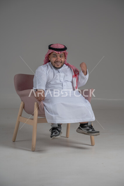 الاناقة والاهتمام بالمظهر الخارجي، بورتريه لرجل عربي خليجي سعودي مبتسم قصير القامة يجلس على كرسي مريح بإيماءات الفرح والسرور والحماس، خلفية رمادية