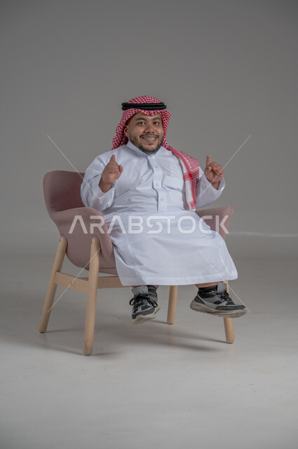 الاناقة والاهتمام بالمظهر الخارجي، بورتريه لرجل عربي خليجي سعودي مبتسم قصير القامة يجلس على كرسي مريح بإيماءات الفرح والسرور والحماس، خلفية رمادية