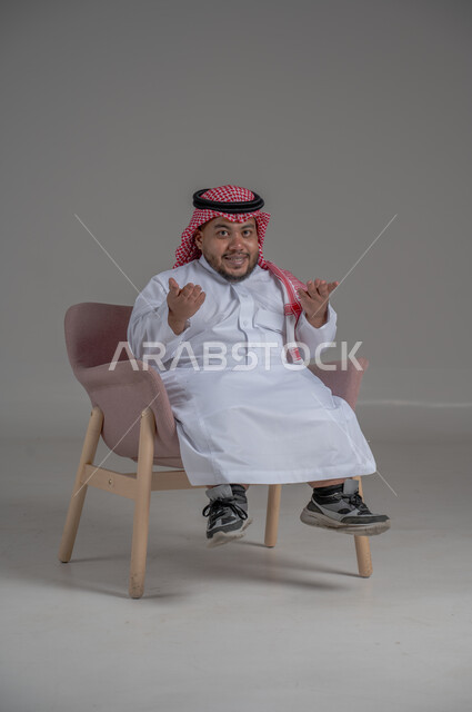 الاناقة والاهتمام بالمظهر الخارجي، بورتريه لرجل عربي خليجي سعودي مبتسم قصير القامة يجلس على كرسي مريح بإيماءات الفرح والسرور والحماس، خلفية رمادية