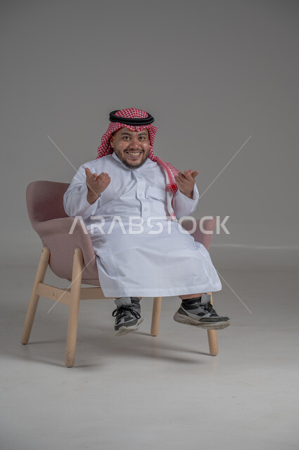 الاناقة والاهتمام بالمظهر الخارجي، بورتريه لرجل عربي خليجي سعودي مبتسم قصير القامة يجلس على كرسي مريح بإيماءات الفرح والسرور والحماس، خلفية رمادية