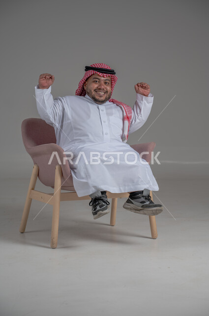 الاناقة والاهتمام بالمظهر الخارجي، بورتريه لرجل عربي خليجي سعودي مبتسم قصير القامة يجلس على كرسي مريح بإيماءات الفرح والسرور والحماس، خلفية رمادية