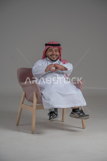 الاناقة والاهتمام بالمظهر الخارجي، بورتريه لرجل عربي خليجي سعودي مبتسم قصير القامة يجلس على كرسي مريح بإيماءات الفرح والسرور والحماس، خلفية رمادية