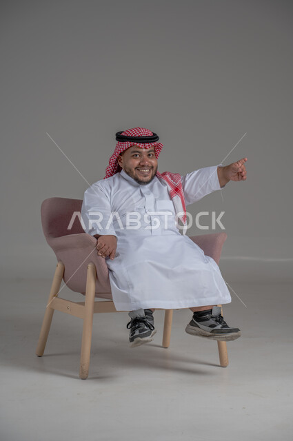 الاناقة والاهتمام بالمظهر الخارجي، بورتريه لرجل عربي خليجي سعودي مبتسم قصير القامة يجلس على كرسي مريح بإيماءات الفرح والسرور والحماس، خلفية رمادية