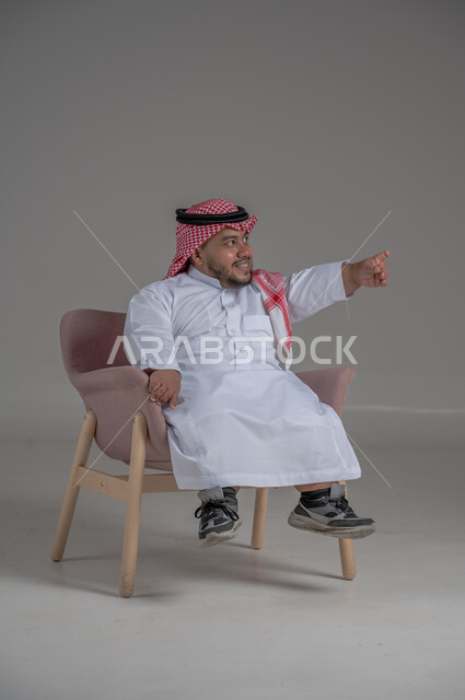 الاناقة والاهتمام بالمظهر الخارجي، بورتريه لرجل عربي خليجي سعودي مبتسم قصير القامة يجلس على كرسي مريح بإيماءات الفرح والسرور والحماس، خلفية رمادية