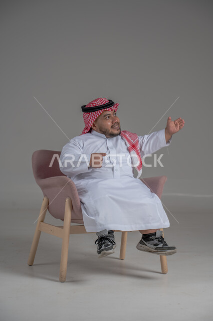 الاناقة والاهتمام بالمظهر الخارجي، بورتريه لرجل عربي خليجي سعودي مبتسم قصير القامة يجلس على كرسي مريح بإيماءات الفرح والسرور والحماس، خلفية رمادية