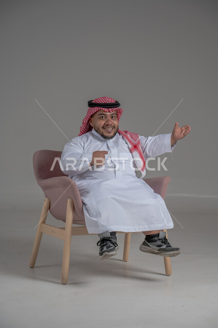 الاناقة والاهتمام بالمظهر الخارجي، بورتريه لرجل عربي خليجي سعودي مبتسم قصير القامة يجلس على كرسي مريح بإيماءات الفرح والسرور والحماس، خلفية رمادية
