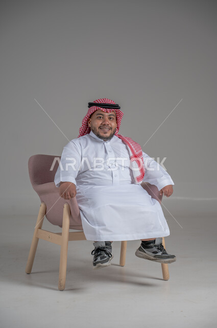 مفهوم الاناقة والاهتمام بالمظهر الخارجي، بورتريه لرجل عربي خليجي سعودي مبتسم قصير القامة يجلس على كرسي مريح بإيماءات الفرح والسرور والحماس، خلفية رمادية