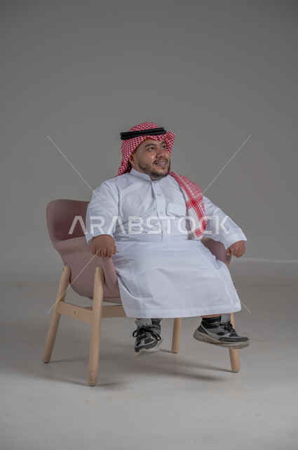 The concept of elegance and interest in external appearance, portrait of a smiling short Saudi Arabian Gulf man sitting on a comfortable chair with gestures of joy, happiness and enthusiasm, gray background