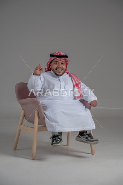 مفهوم الاناقة والاهتمام بالمظهر الخارجي، بورتريه لرجل عربي خليجي سعودي مبتسم قصير القامة يجلس على كرسي مريح بإيماءات الفرح والسرور والحماس، خلفية رمادية