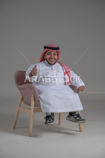 مفهوم الاناقة والاهتمام بالمظهر الخارجي، بورتريه لرجل عربي خليجي سعودي مبتسم قصير القامة يجلس على كرسي مريح بإيماءات الفرح والسرور والحماس، خلفية رمادية