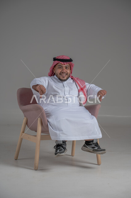 The concept of elegance and interest in external appearance, portrait of a smiling short Saudi Arabian Gulf man sitting on a comfortable chair with gestures of joy, happiness and enthusiasm, gray background