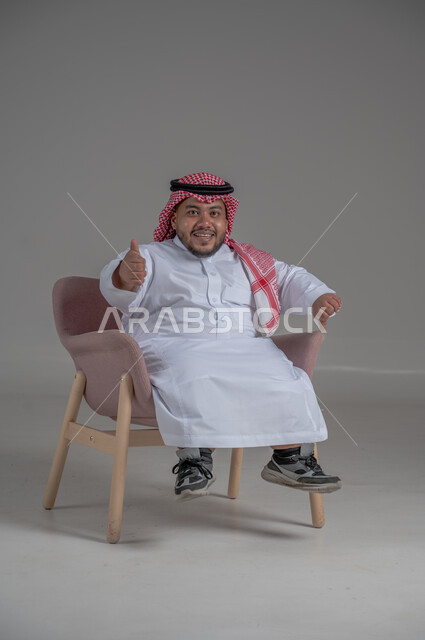 مفهوم الاناقة والاهتمام بالمظهر الخارجي، بورتريه لرجل عربي خليجي سعودي مبتسم قصير القامة يجلس على كرسي مريح بإيماءات الفرح والسرور والحماس، خلفية رمادية