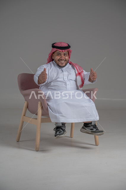 مفهوم الاناقة والاهتمام بالمظهر الخارجي، بورتريه لرجل عربي خليجي سعودي مبتسم قصير القامة يجلس على كرسي مريح بإيماءات الفرح والسرور والحماس، خلفية رمادية