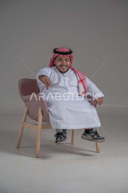 The concept of elegance and interest in external appearance, portrait of a smiling short Saudi Arabian Gulf man sitting on a comfortable chair with gestures of joy, happiness and enthusiasm, gray background