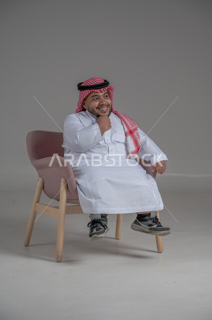 مفهوم الاناقة والاهتمام بالمظهر الخارجي، بورتريه لرجل عربي خليجي سعودي مبتسم قصير القامة يجلس على كرسي مريح بإيماءات الفرح والسرور والحماس، خلفية رمادية