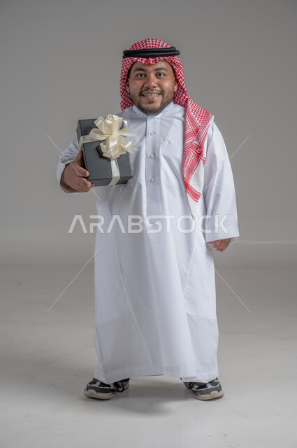 Saudi society support for people with disabilities, happy occasions and holidays, portrait of a disabled Saudi Arabian Gulf man wearing traditional thobe and shemagh sitting on a wheelchair holding a gift box, the feeling of love and appreciation for the disabled, full body image, gray background