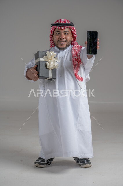 Saudi society support for people with disabilities, happy occasions and holidays, portrait of a Saudi Arabian Gulf dwarf man wearing traditional thobe and shemagh holding a gift box and mobile phone, the feeling of love and appreciation for the disabled, full body image, gray background