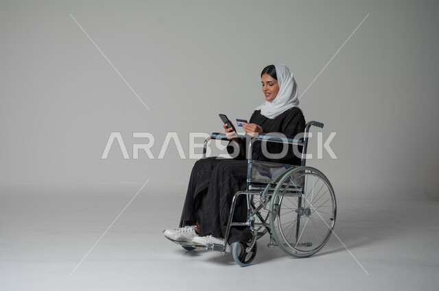استخدام التقنيات الحديثة والمتطورة، بورتريه لإمرأة عربية خليجية سعودية مقعدة من ذوي الهمم ترتدي الحجاب والعباءة السوداء تمسك بيدها الهاتف المحمول وبطاقة الائتمان البنكية تجلس على كرسي متحرك، الشراء والدفع من خلال برامج وتطبيقات الجوال، خلفية رمادية