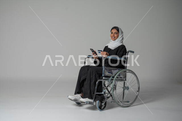 استخدام التقنيات الحديثة والمتطورة، بورتريه لإمرأة عربية خليجية سعودية مقعدة من ذوي الهمم ترتدي الحجاب والعباءة السوداء تمسك بيدها الهاتف المحمول وبطاقة الائتمان البنكية تجلس على كرسي متحرك، الشراء والدفع من خلال برامج وتطبيقات الجوال، خلفية رمادية