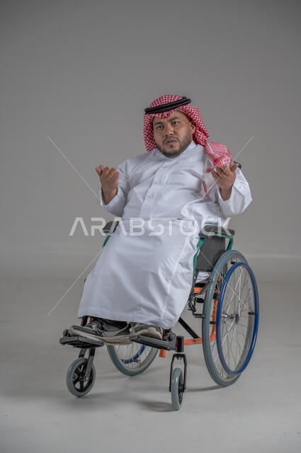 نمط حياة ذوي الهمم، استخدام كرسي المعاقين للمساعدة على الحركة والتنقل، بورتريه لشاب عربي خليجي سعودي قزم من ذوي الهمم يرتدي الثوب التقليدي والشماغ يجلس على الكرسي المتحرك يقوم بتحريك يديه بايماءات الغضب، صورة الجسم بالطول الكامل، خلفية رمادية