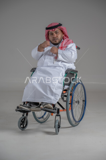 نمط حياة ذوي الهمم، استخدام كرسي المعاقين للمساعدة على الحركة والتنقل، بورتريه لشاب عربي خليجي سعودي قزم من ذوي الهمم يرتدي الثوب التقليدي والشماغ يجلس على الكرسي المتحرك يقوم بتحريك يديه بايماءات الغضب، صورة الجسم بالطول الكامل، خلفية رمادية