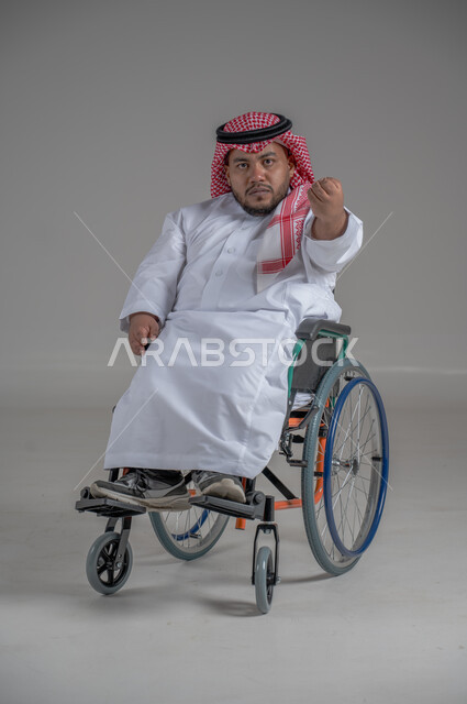 نمط حياة ذوي الهمم، استخدام كرسي المعاقين للمساعدة على الحركة والتنقل، بورتريه لشاب عربي خليجي سعودي قزم من ذوي الهمم يرتدي الثوب التقليدي والشماغ يجلس على الكرسي المتحرك يقوم بتحريك يديه بايماءات الغضب، صورة الجسم بالطول الكامل، خلفية رمادية