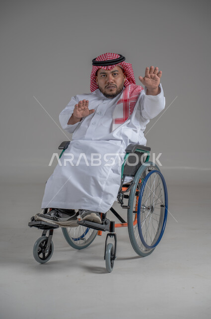 نمط حياة ذوي الهمم، استخدام كرسي المعاقين للمساعدة على الحركة والتنقل، بورتريه لشاب عربي خليجي سعودي قزم من ذوي الهمم يرتدي الثوب التقليدي والشماغ يجلس على الكرسي المتحرك يقوم بتحريك يديه بايماءات التوقف عن فعل شيء، صورة الجسم بالطول الكامل، خلفية رمادية