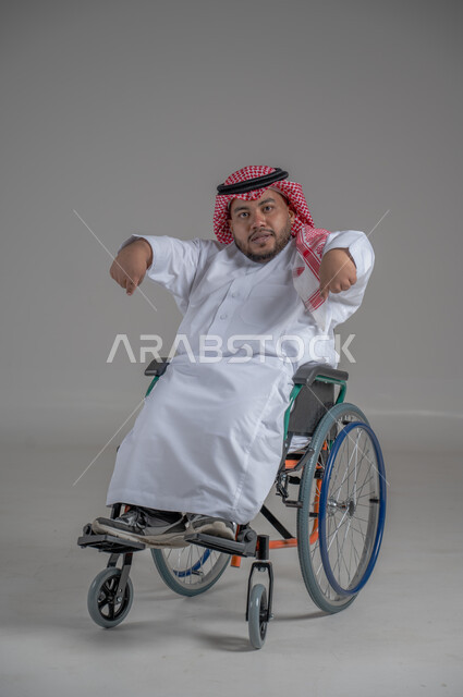 نمط حياة ذوي الهمم، استخدام كرسي المعاقين للمساعدة على الحركة والتنقل، بورتريه لشاب عربي خليجي سعودي قزم من ذوي الهمم يرتدي الثوب التقليدي والشماغ يجلس على الكرسي المتحرك يقوم بتحريك يديه بايماءات السرور، صورة الجسم بالطول الكامل، خلفية رمادية