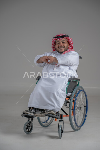 Lifestyle of people with disabilities, using a wheelchair to assist with mobility and transportation, portrait of a young Saudi Arabian Gulf dwarf with disabilities wearing traditional thobe and shemagh sitting on a wheelchair moving his hands with gestures of pleasure, full body image, gray background
