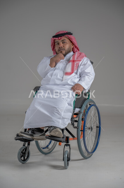 نمط حياة ذوي الهمم، استخدام كرسي المعاقين للمساعدة على الحركة والتنقل، بورتريه لشاب عربي خليجي سعودي قزم من ذوي الهمم يرتدي الثوب التقليدي والشماغ يجلس على الكرسي المتحرك يقوم بتحريك يديه بايماءات السرور، صورة الجسم بالطول الكامل، خلفية رمادية