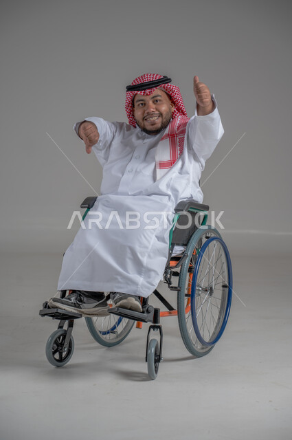 نمط حياة ذوي الهمم، استخدام كرسي المعاقين للمساعدة على الحركة والتنقل، بورتريه لشاب عربي خليجي سعودي قزم من ذوي الهمم يرتدي الثوب التقليدي والشماغ يجلس على الكرسي المتحرك يقوم بتحريك يديه بايماءات السرور، صورة الجسم بالطول الكامل، خلفية رمادية