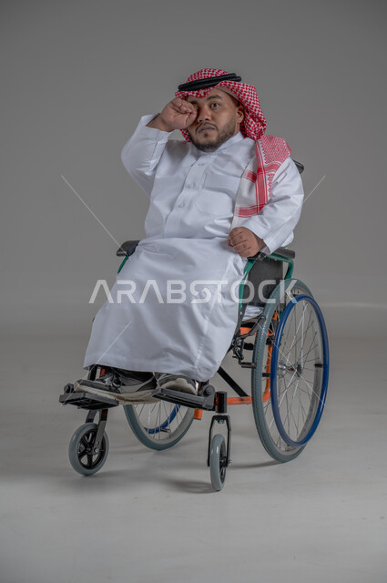 استخدام كرسي المعاقين للمساعدة على الحركة والتنقل، نمط حياة ذوي الهمم، بورتريه لشاب عربي خليجي سعودي قزم من ذوي الهمم يرتدي الثوب التقليدي والشماغ يجلس على الكرسي المتحرك يقوم بتحريك يديه بايماءات السعادة، صورة الجسم بالطول الكامل، خلفية رمادية
