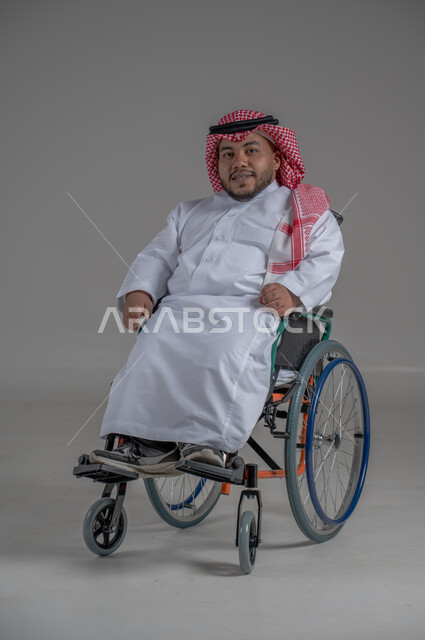استخدام كرسي المعاقين للمساعدة على الحركة والتنقل، نمط حياة ذوي الهمم، بورتريه لشاب عربي خليجي سعودي قزم من ذوي الهمم يرتدي الثوب التقليدي والشماغ يجلس على الكرسي المتحرك يقوم بتحريك يديه بايماءات السعادة، صورة الجسم بالطول الكامل، خلفية رمادية