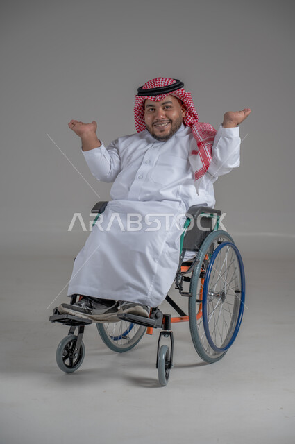 استخدام كرسي المعاقين للمساعدة على الحركة والتنقل، نمط حياة ذوي الهمم، بورتريه لشاب عربي خليجي سعودي قزم من ذوي الهمم يرتدي الثوب التقليدي والشماغ يجلس على الكرسي المتحرك يقوم بتحريك يديه بايماءات السعادة، صورة الجسم بالطول الكامل، خلفية رمادية