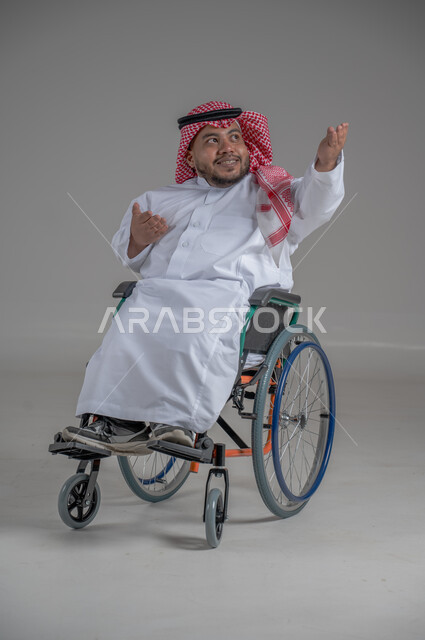 استخدام كرسي المعاقين للمساعدة على الحركة والتنقل، نمط حياة ذوي الهمم، بورتريه لشاب عربي خليجي سعودي قزم من ذوي الهمم يرتدي الثوب التقليدي والشماغ يجلس على الكرسي المتحرك يقوم بتحريك يديه بايماءات السعادة، صورة الجسم بالطول الكامل، خلفية رمادية