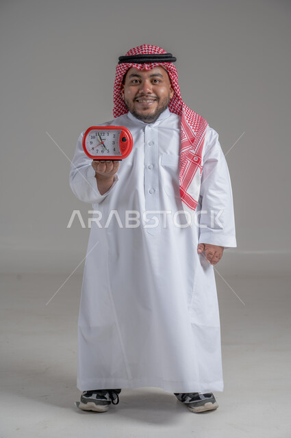 Timer, concept of masculinity and self-confidence, portrait of a Saudi Arabian Gulf dwarf man wearing traditional thobe and shemagh holding a small watch looking at the camera with an expression of pleasure, standing straight and steady, full body image, gray background