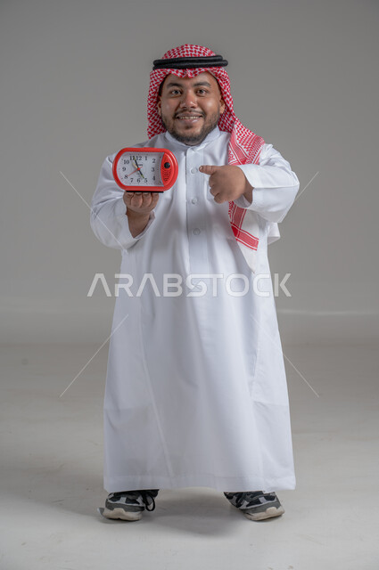 Timer, concept of masculinity and self-confidence, portrait of a Saudi Arabian Gulf dwarf man wearing traditional thobe and shemagh holding a small watch looking at the camera with an expression of pleasure, standing straight and steady, full body image, gray background