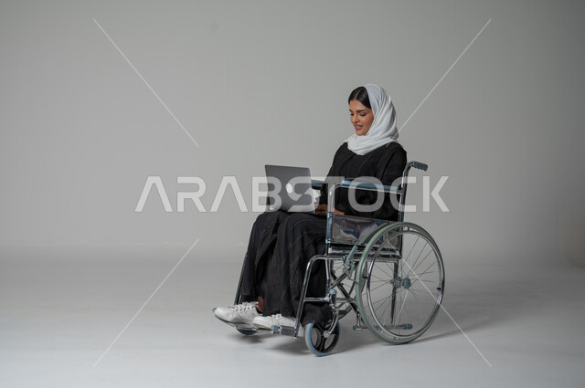 العمل عن بعد وتصفح الانترنت، استخدام التكنولوجيا الحديثة وانجازالمهام، بورتريه مقرب لامرأة عربية سعودية خليجية من ذوي الاحتياجات الخاصة ترتدي العباءة السوداء والحجاب تجلس على كرسي متحرك تعمل على الحاسب المحمول، خلفية رمادية