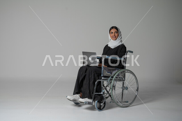العمل عن بعد وتصفح الانترنت، استخدام التكنولوجيا الحديثة وانجازالمهام، بورتريه مقرب لامرأة عربية سعودية خليجية من ذوي الاحتياجات الخاصة ترتدي العباءة السوداء والحجاب تجلس على كرسي متحرك تمسك بيدها حاسب محمول تنظر إلى شيء ما، خلفية رمادية