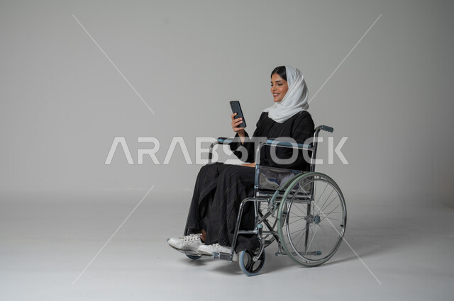 إيماءات وتعابير وجه تدل على السعادة بإجراء مكالمة فيديو, استخدام تطبيقات الهاتف المحمول، بورتريه مقرب لامرأة عربية سعودية خليجية من ذوي الاحتياجات الخاصة ترتدي العباءة السوداء والحجاب تجلس على كرسي متحرك تمسك بيدها الهاتف المحمول، خلفية رمادية