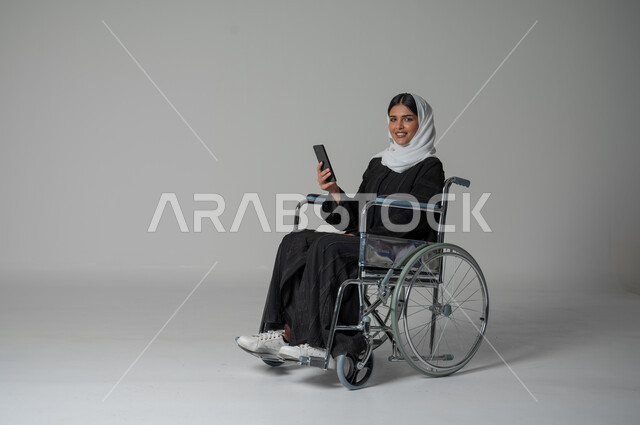 إيماءات وتعابير وجه تدل على السعادة بإجراء مكالمة فيديو, استخدام تطبيقات الهاتف المحمول، بورتريه مقرب لامرأة عربية سعودية خليجية من ذوي الاحتياجات الخاصة ترتدي العباءة السوداء والحجاب تجلس على كرسي متحرك تمسك بيدها الهاتف المحمول، خلفية رمادية