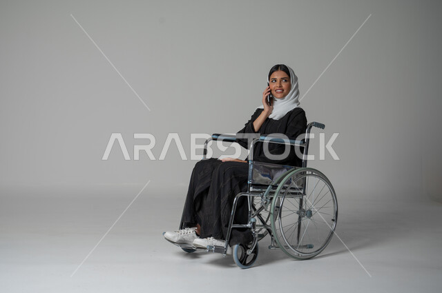 التواصل والاطمئنان على الأصدقاء، بورتريه مقرب لامرأة عربية سعودية خليجية من ذوي الاحتياجات الخاصة ترتدي العباءة السوداء والحجاب تجلس على كرسي متحرك تقوم بإجراء مكالمة صوتية عبر الجوال، الإجابة والرد على الاتصالات بإستخدام الهاتف المحمول، خلفية رمادية